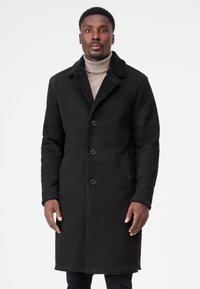 Sort ruskindfrakke med en shearling-krave, der har knaplukning og to frontlommer, glat tekstur og knælængde design.