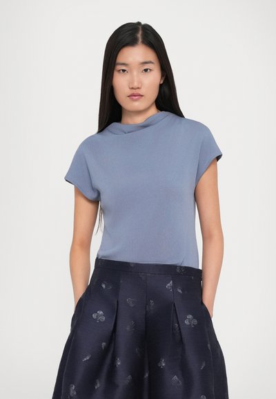 Young woman with long black hair wearing a short-sleeve blue top and high-waisted dark skirt with subtle patterns, hands in pockets, neutral background.