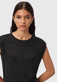 Una giovane donna con lunghi capelli scuri indossa un top senza maniche nero a pois e piccoli orecchini quadrati d'oro su uno sfondo chiaro e neutro.