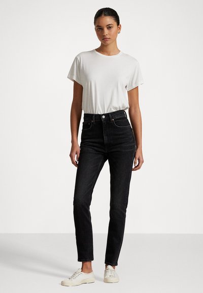 Mujer de pie vistiendo una camiseta blanca de manga corta metida dentro de unos pantalones vaqueros negros de cintura alta y zapatillas blancas, contra un fondo liso.