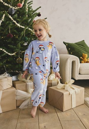 Laps pidulikes pidžaamades naeratamas jõuluehtes kuusepuu lähedal, ümbritsetud kingitustega puidust põrandal hubases elutoas.