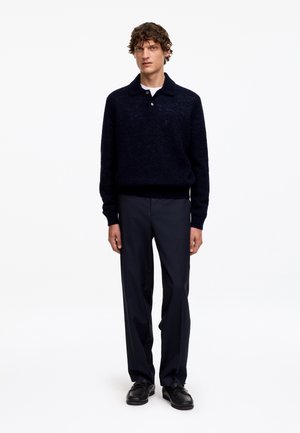 Homme portant un pull bleu marine foncé avec col, pantalon droit foncé et chaussures noires, debout devant un fond blanc uni.