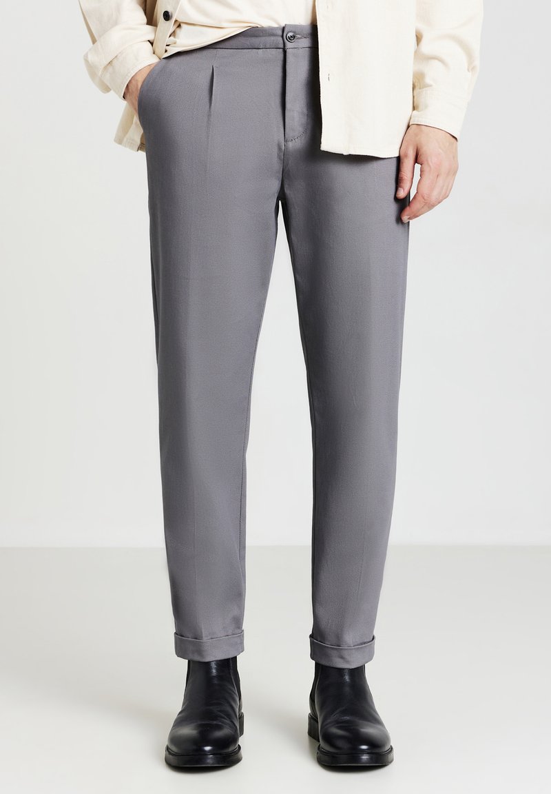 Pantaloni grigi su misura con una texture liscia, davanti plissettato e orli arrotolati, abbinati a stivali in pelle nera.