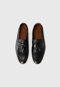 Mocassins en cuir noir avec des glands, dotés de semelles intérieures marron, disposés de manière symétrique sur un fond blanc.