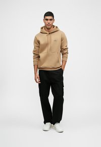 Άνδρας που στέκεται φορώντας καφέ φούτερ με κουκούλα, μαύρο cargo παντελόνι και λευκά αθλητικά παπούτσια, μπροστά από απλή φωτεινή φόντο.