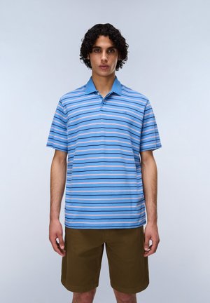 Nuori mies kiharatukkaisena, sinivalkoraidallisessa ja vaaleanpunaisessa poolopaidassa sekä ruskeissa shortseissa, seisoo yksiväristä taustaa vasten.