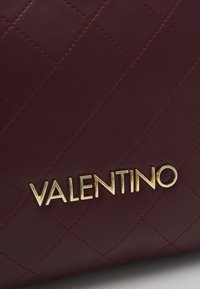 Burgundska usnjena torba s prešito zasnovo, opremljena z zlatim logotipom "VALENTINO". Vidne so podrobne šive po celotnem materialu.
