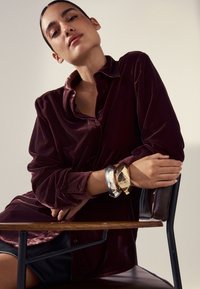 Bordeauxrotes Samthemd mit Kragen, getragen über einem schwarzen Lederrock. Drei klobige metallische Armbänder in Gold, Silber und Bronze am Handgelenk.