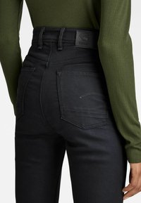 Černé džínsy s vysokým pasem, hladkým povrchem, s ozdobnými zadními kapsami a značkovou černou koženou nášivkou na pase.