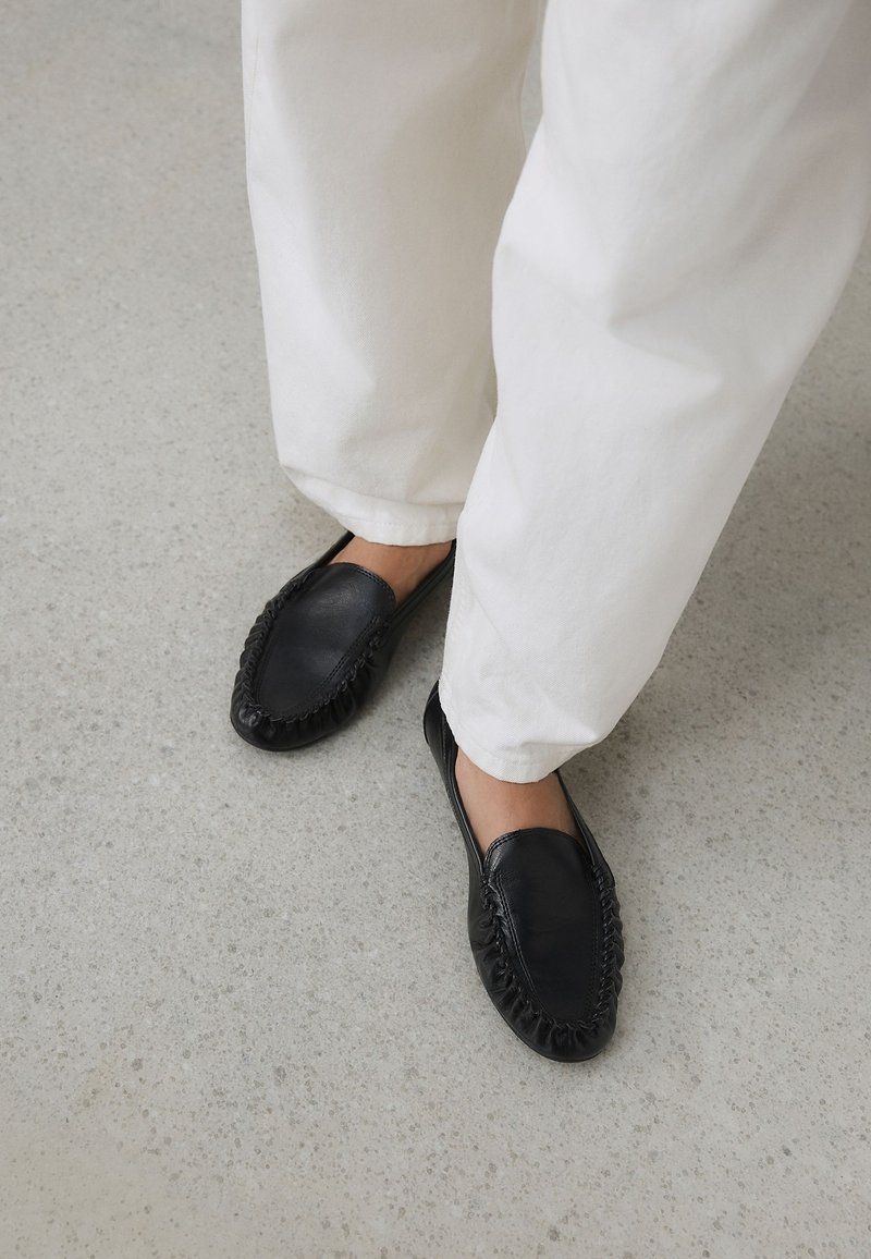 Personne portant un pantalon blanc et des chaussures à enfiler en cuir noir style mocassin, debout sur un sol gris clair.