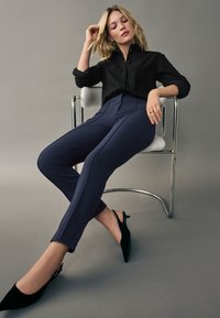 Cămașă neagră cu dungi, pantaloni navy cu talie înaltă și pantofi cu toc slingback negri, așezată pe un scaun din crom pe un fundal gri.