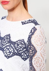 Top in pizzo con un corpo bianco e dettagli in pizzo floreale blu navy sulle spalle e sulle maniche, che mostrano motivi e trame intricate.