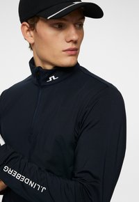 Maglietta sportiva a maniche lunghe blu navy con zip e colletto alto, caratterizzata da un logo bianco sulla spalla sinistra e dettagli testuali lungo la manica.