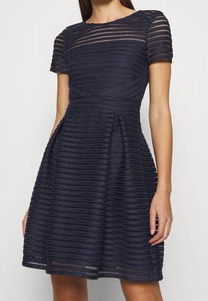 Femme portant une robe bleu marine mi-genou avec des rayures horizontales en dentelle, des manches courtes, une taille ajustée et une jupe plissée.