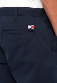 Ναυτικά μπλε παντελόνια με λεία υφή, διαθέτοντας μια πίσω τσέπη με ένα λογότυπο "TOMMY JEANS" σε κόκκινα, λευκά και μπλε χρώματα.