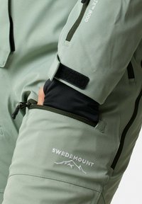 Jasnozielona kurtka outdoorowa z zamkniętą na zamek kieszenią, mankietami na rzepy oraz białym haftowanym logo "SWEDEMOUNT". Wykonana z wodoodpornego materiału.