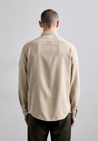 Beige puuvillainen pitkähiha-paita, jossa on kaareva helma, napeilla suljettavat kalvosimet ja kaulus. Yksinkertainen muotoilu ja sileä pinta, esitetty takaapäin.