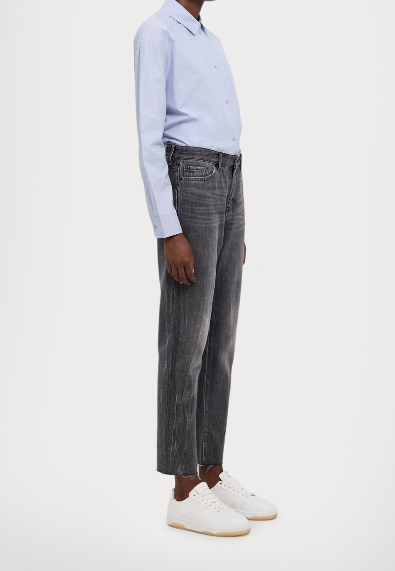 Personne portant une chemise à manches longues bleu clair, un jean noir délavé à jambes droites avec des ourlets effilochés, et des baskets blanches, debout sur un fond uni.