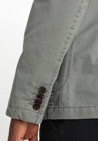 Zbliżenie na rękaw zielonej kurtki z czterema brązowymi guzikami i widocznym szwem wzdłuż mankietu i szwu bocznego.