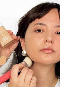 Botella de base en color marrón sostenida en una mano, aplicando con un pincel en la otra; la modelo tiene pendientes de plata y piel suave y saludable.