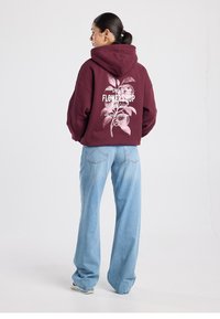 Viininpunainen hupullinen paita, jossa on rento istuvuus ja valkoinen kukkakuvio takana. Yhdistetty vaaleansinisiin leveälahkeisiin farkkuihin.