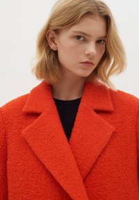 Röd texturerad kappa med notch-krage och breda slag, med en grov väv. Bärs över en svart topp, med en enkel, ren halsringning.