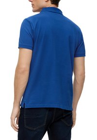 Polo shirt in cotone blu con maniche corte, colletto e spacchi laterali all'orlo, visto da dietro su un modello che indossa jeans scuri.