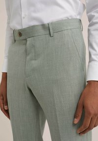 Šviesiai žalios kelnės su tekstūruotu raštu, turinčios sagčių uždarymą ir diržų kilpas, derinamos su baltu marškiniu su sagutėmis.