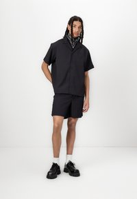 Kortærmet, knap-åben skjorte og matchende shorts i mørk stof med subtile lodrette striber. Båret med sorte sko og et mønstret tørklæde.