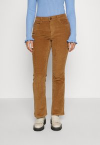 Žena na sobě má hnědé kordové kalhoty, světle modrý úplet s dlouhým rukávem a volánkovými manžetami a krémové loafers s tlustou podrážkou.