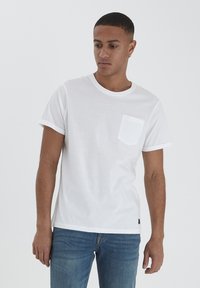 T-shirt bianca in cotone con maniche corte, scollatura rotonda e taschino sul lato sinistro del petto. Indossata con jeans in denim blu. Design minimalista.