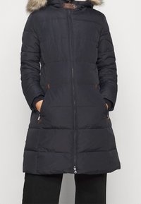 Marinblå pufferjacka med avtagbar huva fodrad med päls, dragkedja och sidofickor. Har bruna detaljer och en quiltad textur.