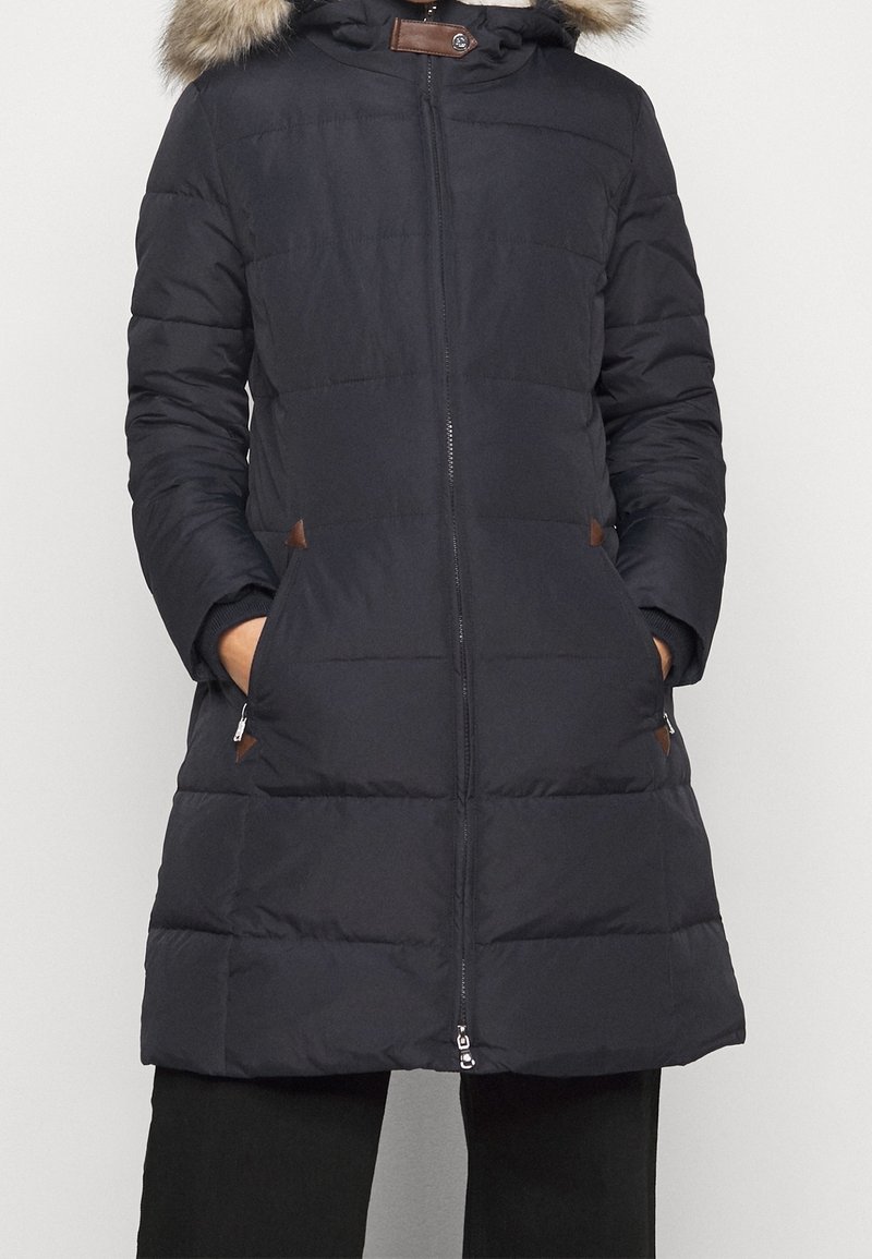 Marinblå pufferjacka med avtagbar huva fodrad med päls, dragkedja och sidofickor. Har bruna detaljer och en quiltad textur.