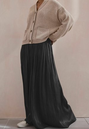 Osoba ubrana w beżowy dziergany kardigan, czarną plisowaną spódnicę maxi oraz białe trampki, stojąca w pomieszczeniu na tle gładkiej ściany.