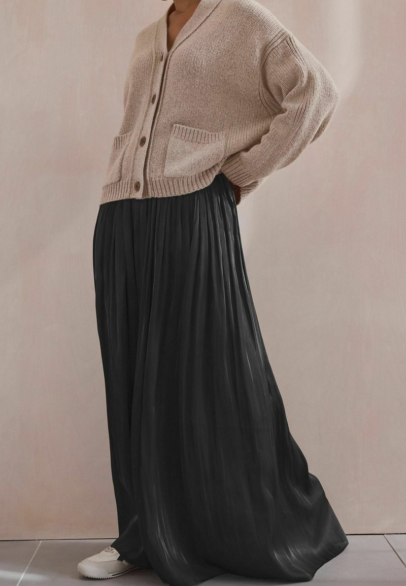 Personne portant un cardigan en maille beige sur une jupe longue plissée noire et des baskets blanches, debout à l'intérieur contre un mur uni.