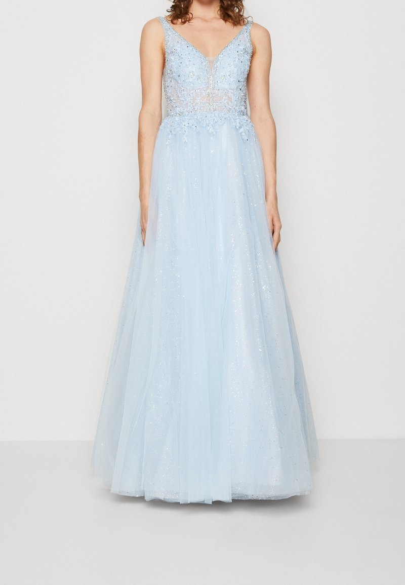 Femme portant une robe sans manches bleu clair avec un décolleté en V profond et un corsage orné, debout devant un fond blanc uni.