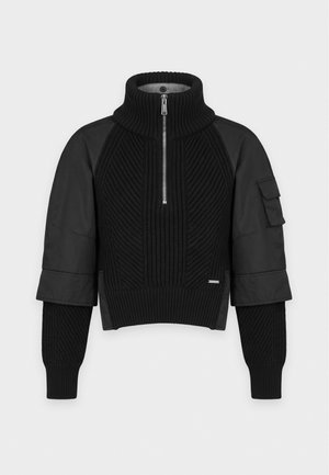 Femme portant un pull noir zippé, une casquette noire, un pantalon foncé avec de grandes poches grises, des talons hauts noirs et tenant un sac à main noir.