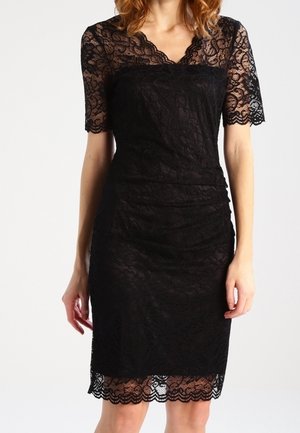 Mujer con vestido negro de encaje hasta la rodilla, mangas cortas, escote con borde ondulado y silueta ajustada, sobre un fondo liso.