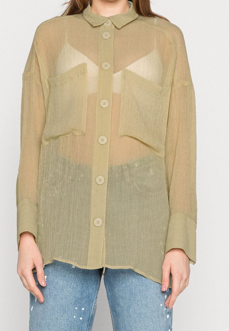Personne portant une chemise boutonnée vert olive transparente avec deux poches avant sur un soutien-gorge beige et un jean bleu avec des éclaboussures de peinture blanches.