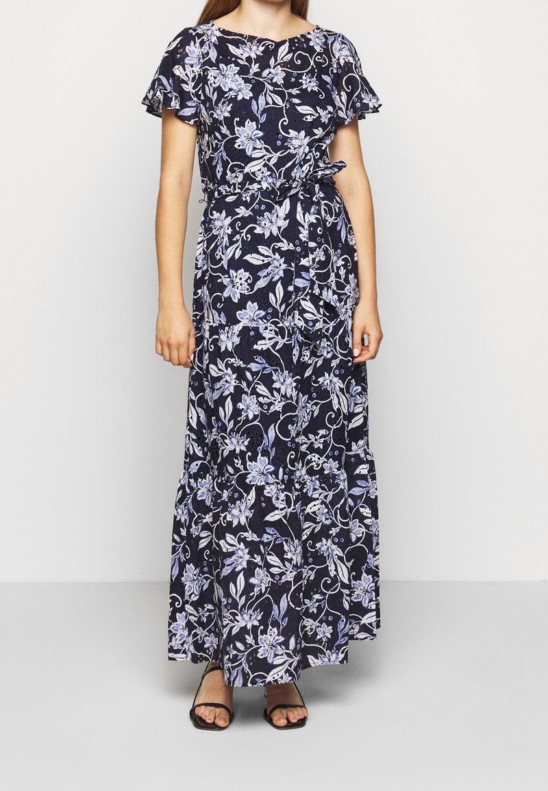 Kvinna iklädd en marinblå blommig maxiklänning med fladdrande ärmar och midjeband, kombinerad med svarta öppna sandaler.