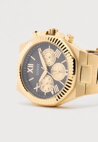 Osmerokotna ura zlato barve z temnosivo številčnico, rimskimi številkami, tremi podštevilčnicami in teksturiranim paščkom. Vidno ime blagovne znamke.