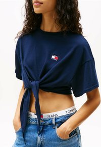 Marineblauw cropped t-shirt met korte mouwen en een geknoopt voorpand, voorzien van een klein logo-embleem. Gedragen met hoge taille blauwe denim jeans.