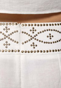 Tejido de prenda blanca con remaches de bronce decorativos en un patrón a lo largo de la cintura, con cierre de cremallera y una textura suave.