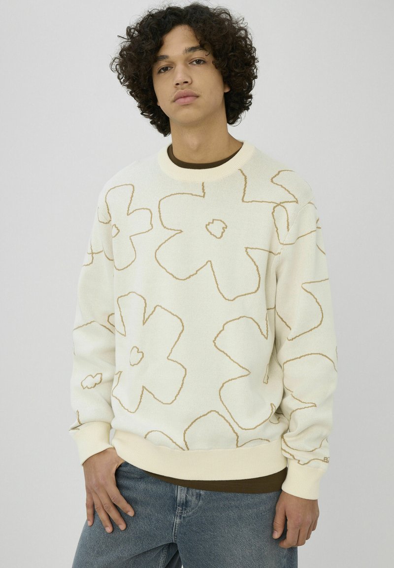 Sweat-shirt crème avec motif floral crème et or, poignets et col côtelés. Porté sur un sous-vêtement marron, assorti à un jean bleu.