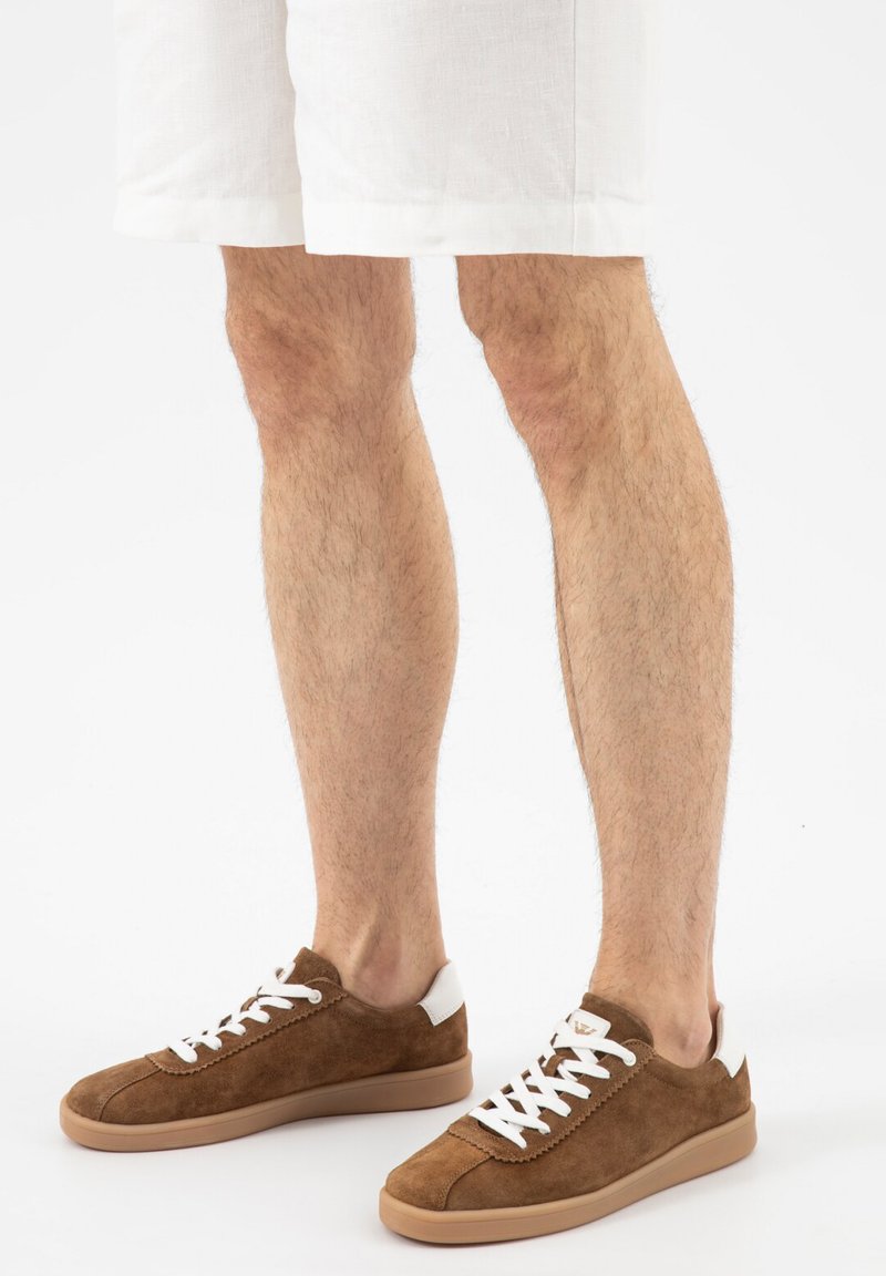 Benen die witte korte broek en bruine suède sneakers met witte veters en lichtbruine zolen dragen, staand op een witte ondergrond.