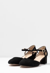 Zapatos de tacón de ante negro con un diseño de tiras en T, punta afilada y un bajo tacón cuadrado. Presentan un accesorio de hebilla dorada y una plantilla beige suave.