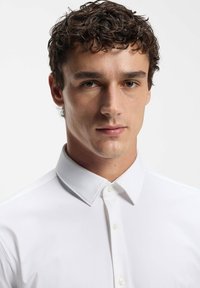 Jonge man met kort, krullend bruin haar, gekleed in een dichtgeknoopt wit overhemd, kijkt recht voor zich uit met een neutrale uitdrukking.