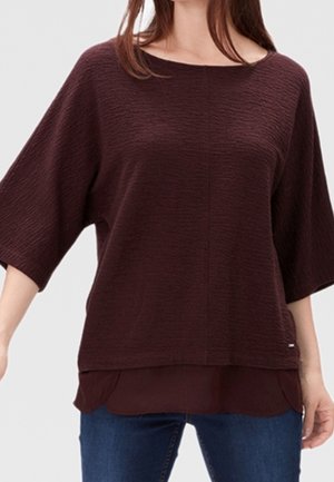 Kvinde iført løs, struktureret mørkebrun overdel med trekvartlange ærmer over en lagdelt skjorte med kant, kombineret med blå jeans.