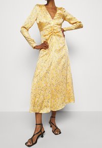 Gul midi-klänning med V-ringning, långa ärmar, rynkad fram och ett vertikalt strukturerat mönster. Matchas med svarta remsandaler med klack.