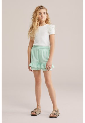 Dekle z dolgimi kodrastimi lasmi, oblečeno v belo bluzo z nabranimi rokavi, mint zeleno krilo z nabranim robom, gleženjsko zapestnico in bež sandale, stoji.